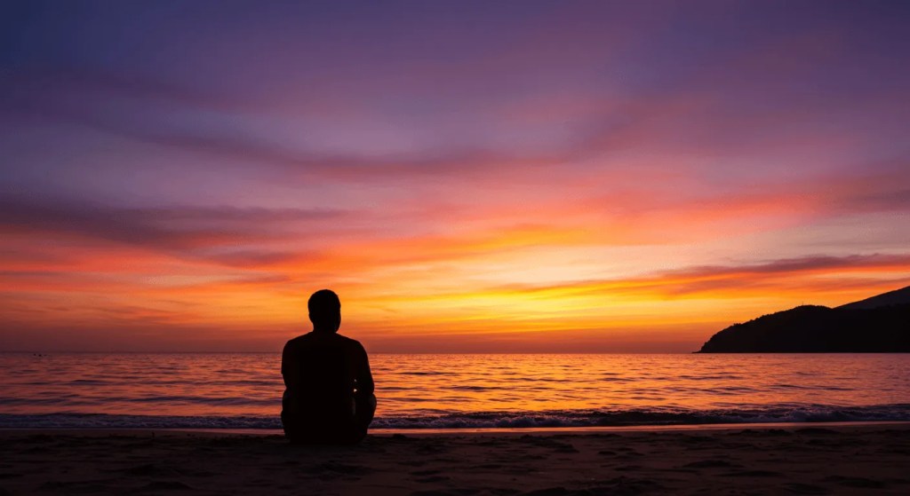 Silhouette of a solo traveler sitting peacefully on a beach, watching a vibrant sunset over the ocean in Southeast Asia.







