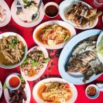 Overhead of a Southeast Asian street-food breakfast spread—fried fish, papaya salad, grilled chicken, soups and sauces on a red table.