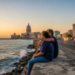Sunset over Havana seen from the Malecón, with locals on the seawall, vintage car lights and the skyline glowing near El Morro.